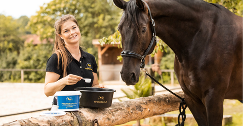 Eine Frau lächelt, während sie Pferdefutter aus einem Behälter zubereitet und neben einem dunkelbraunen Pferd im Freien steht.